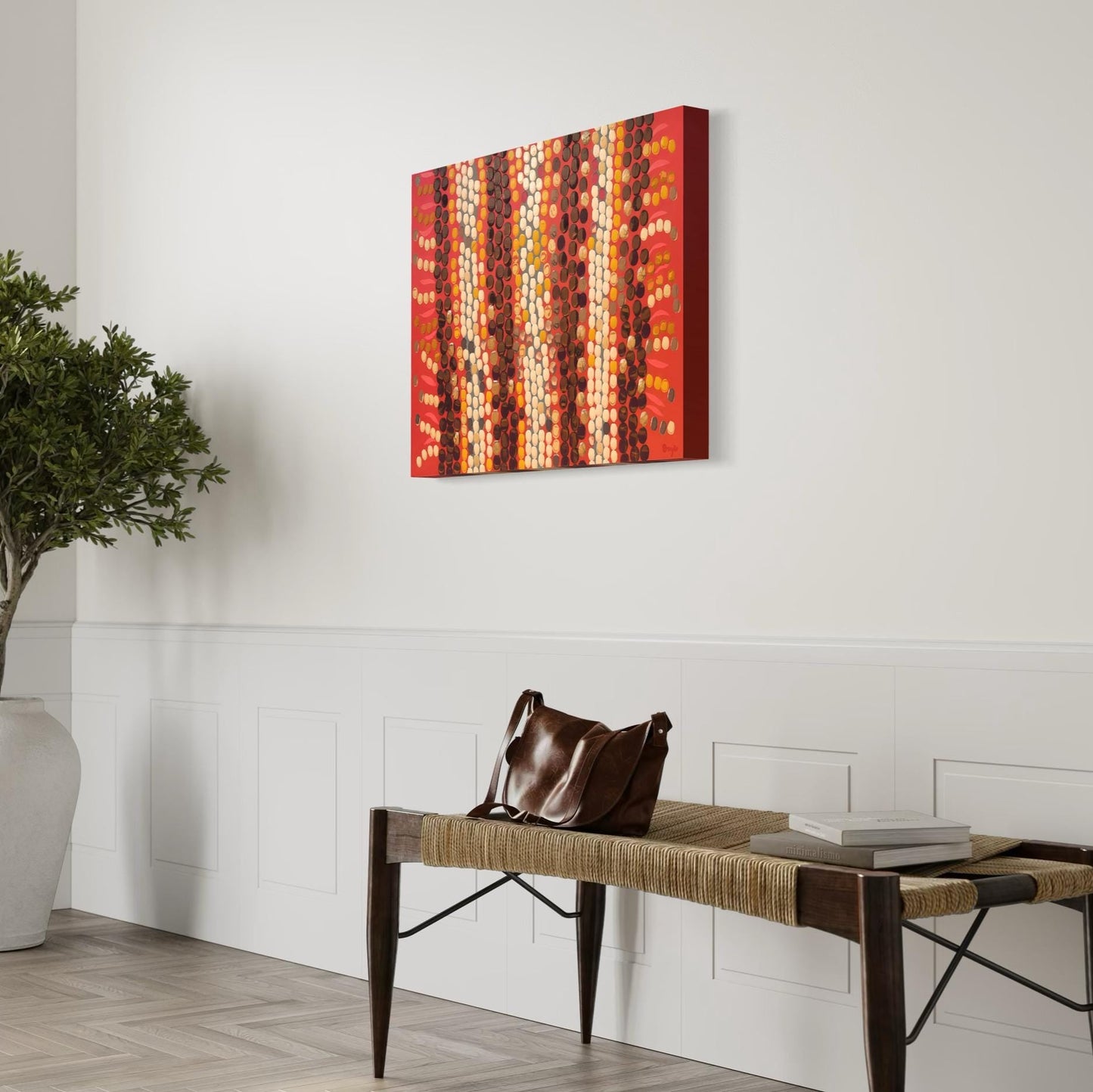 Horizontal Aboriginal dot painting in warm reds, ochres, and browns, side-mounted in a bright entryway above a woven bench with a leather bag, books, and a potted tree adding natural elements.