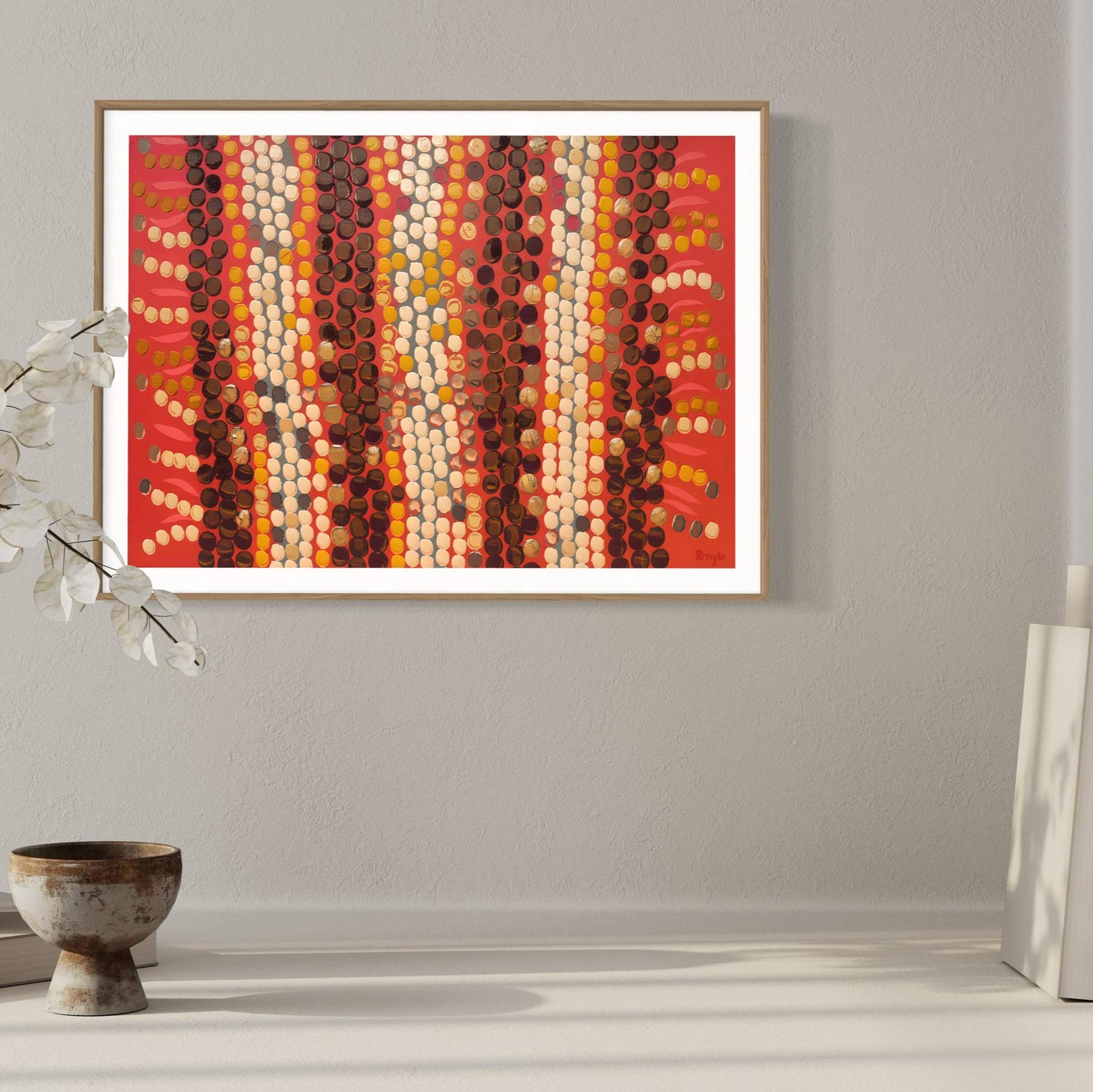 Horizontal Aboriginal dot painting in rich reds, browns, and creams, framed in light wood and displayed on a soft grey wall with neutral ceramic decor and dried white foliage.