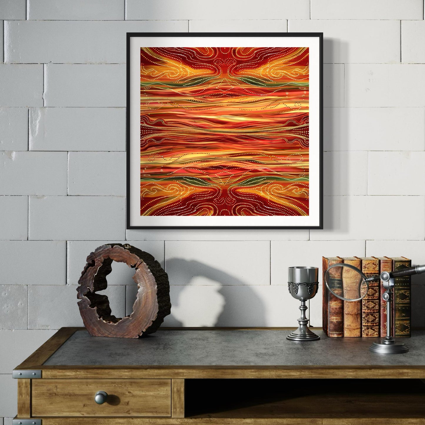 Yurala Flames Aboriginal dot painting printable in fiery earth tones, framed above a rustic wooden desk with vintage books, magnifying glass, and metal goblet against a white brick wall.