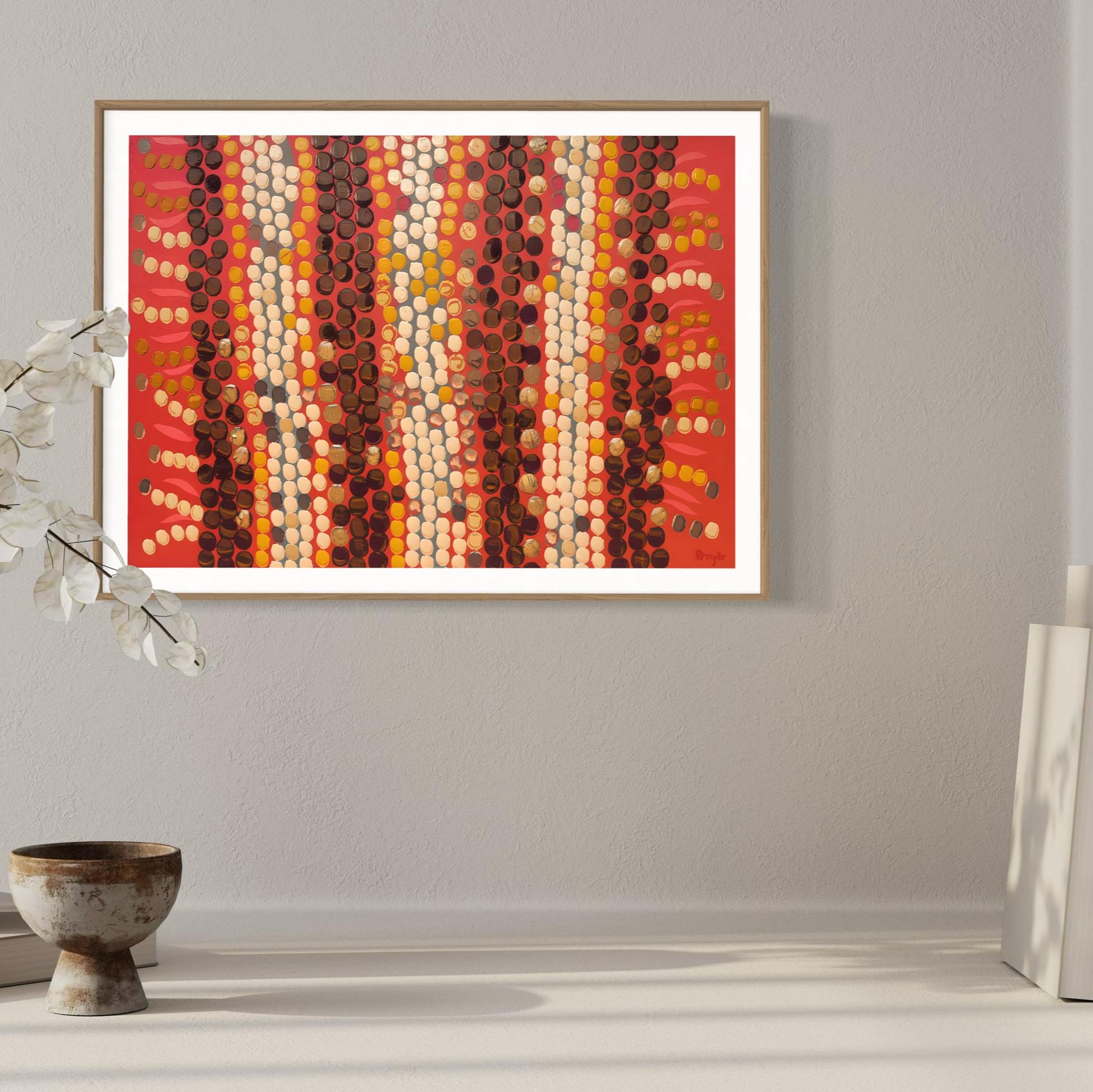 Horizontal Aboriginal dot painting in rich reds, browns, and creams, framed in light wood and displayed on a soft grey wall with neutral ceramic decor and dried white foliage.